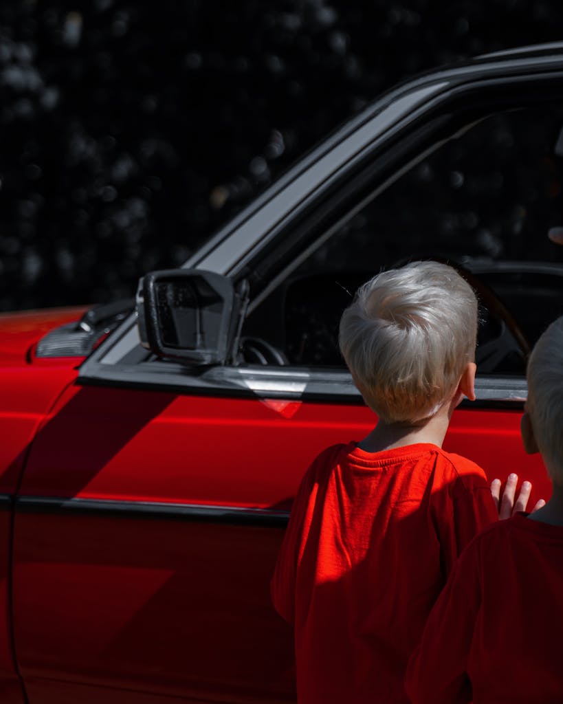 Two blonde boys in red shirts admiring a classic red car, mid-day outdoors.